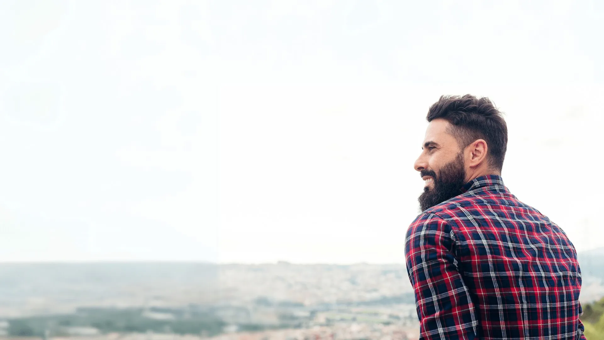 A smiling man with a beard and plaid shirt gazing over a cityscape